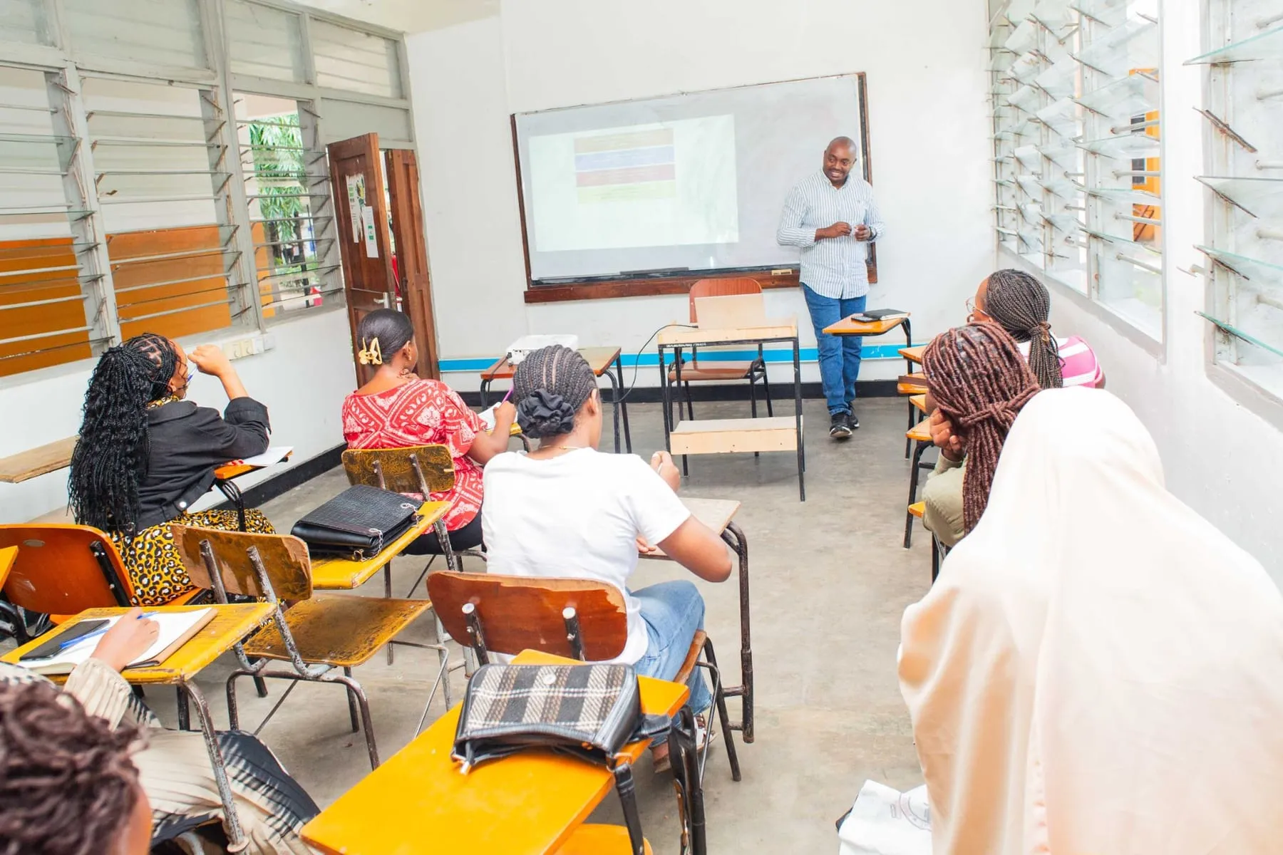 A trainer at the whiteboard presenting slides to students seated at desks in a classroom at the University of Dar es Salaam School of PR & Advertising
